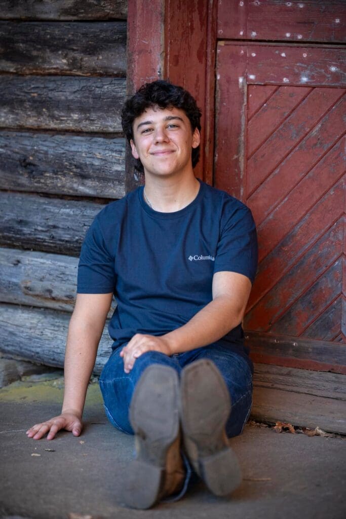 A young man sitting on the ground in front of a log cabin.